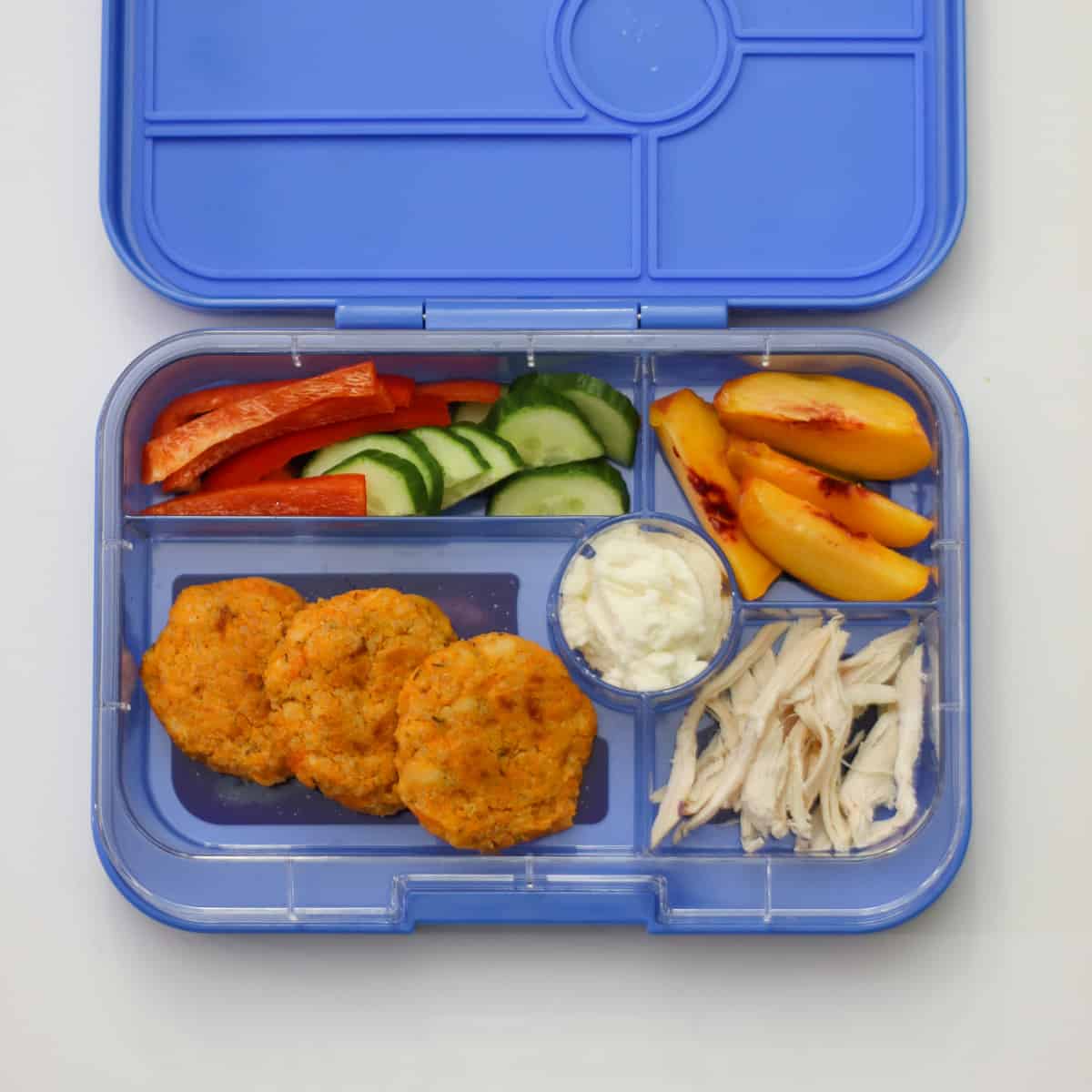 Patties in a school lunch box with ricotta, peaces, cucumber, and bell peppers.