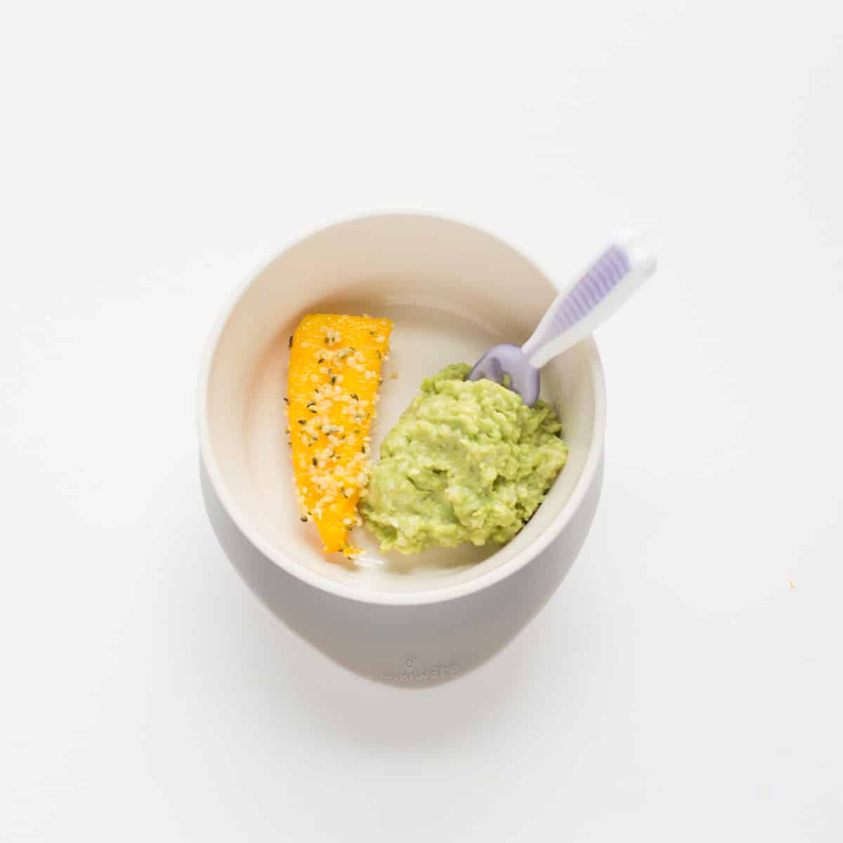 mashed avocado and quinoa with a big strip of mango in a baby bowl with a spoon.