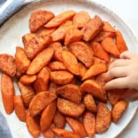 An overhead shot of roasted orange vegetables with toddler's hand reaching for one.