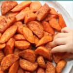 An overhead shot of roasted orange vegetables with toddler's hand reaching for one.