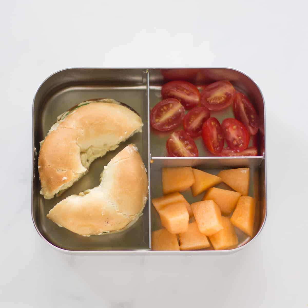 Mini bagel sandwich, cantaloupe, and cherry tomatoes in a stainless steel lunch box.