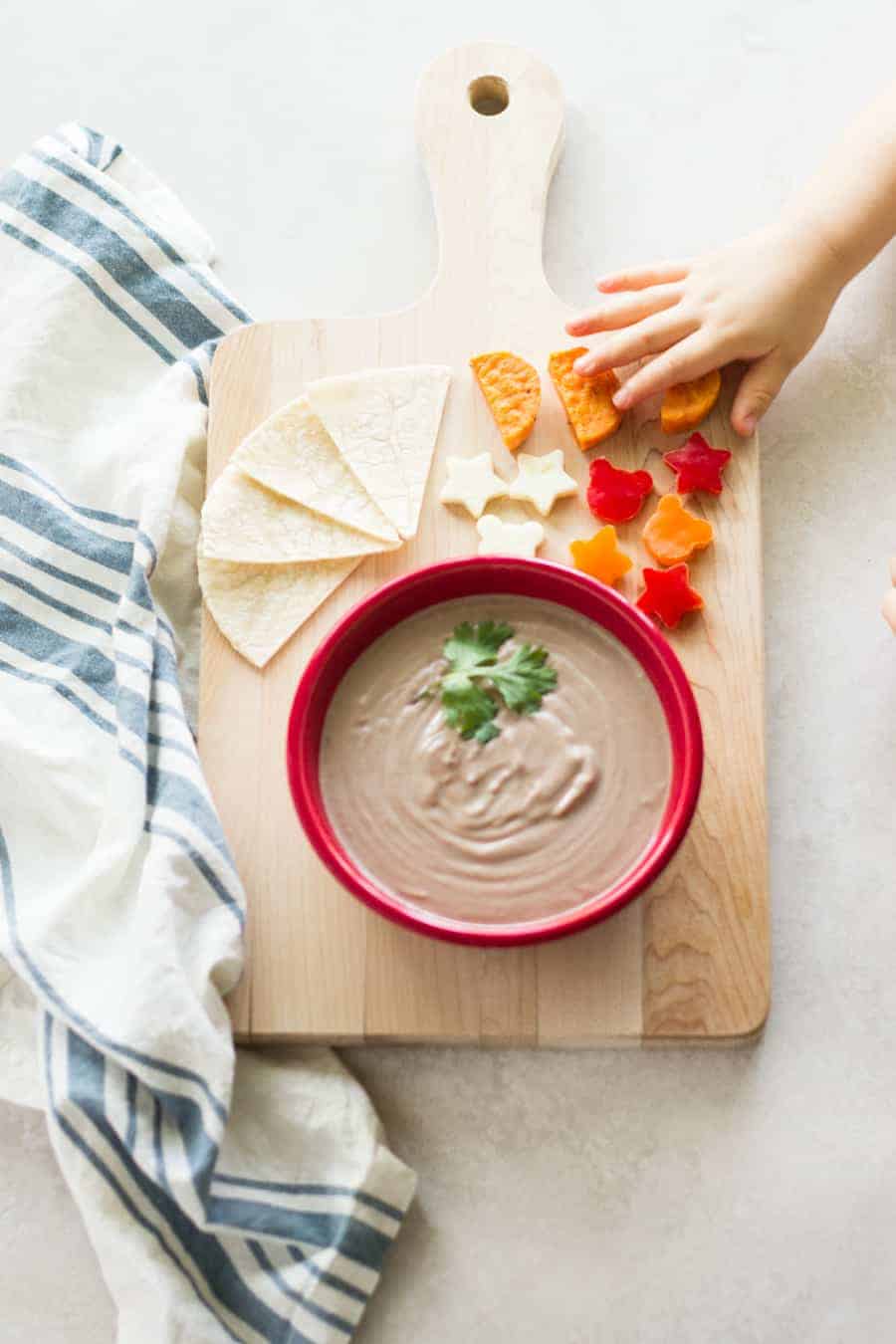 yogurt black bean dip served in a red bowl with tortilla, fun-shaped vegetables and sweet potatoes all placed on a wooden board