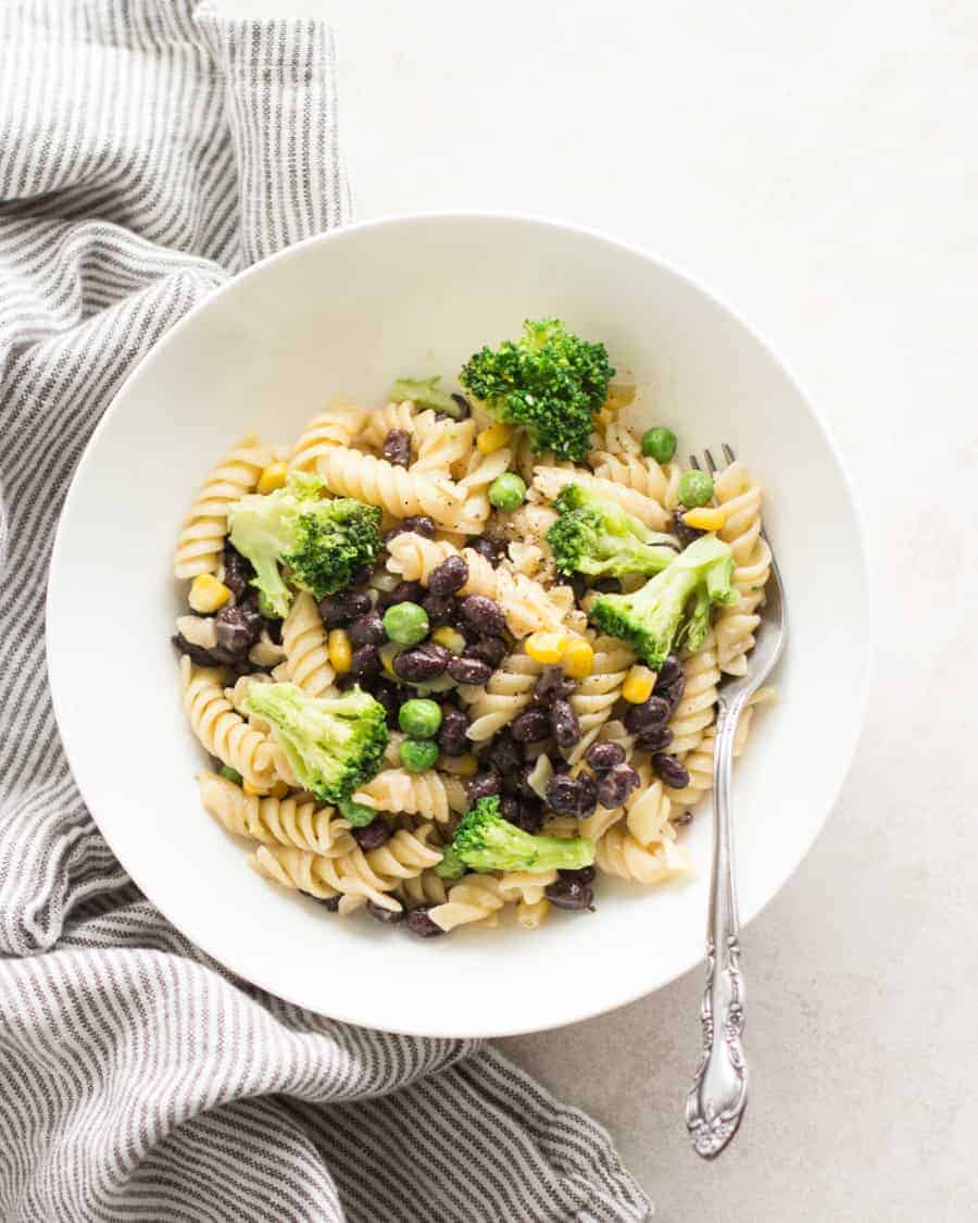 a big bowl of buttered pasta with broccoli, black beans, and peas with a spoon and gray towel on the side