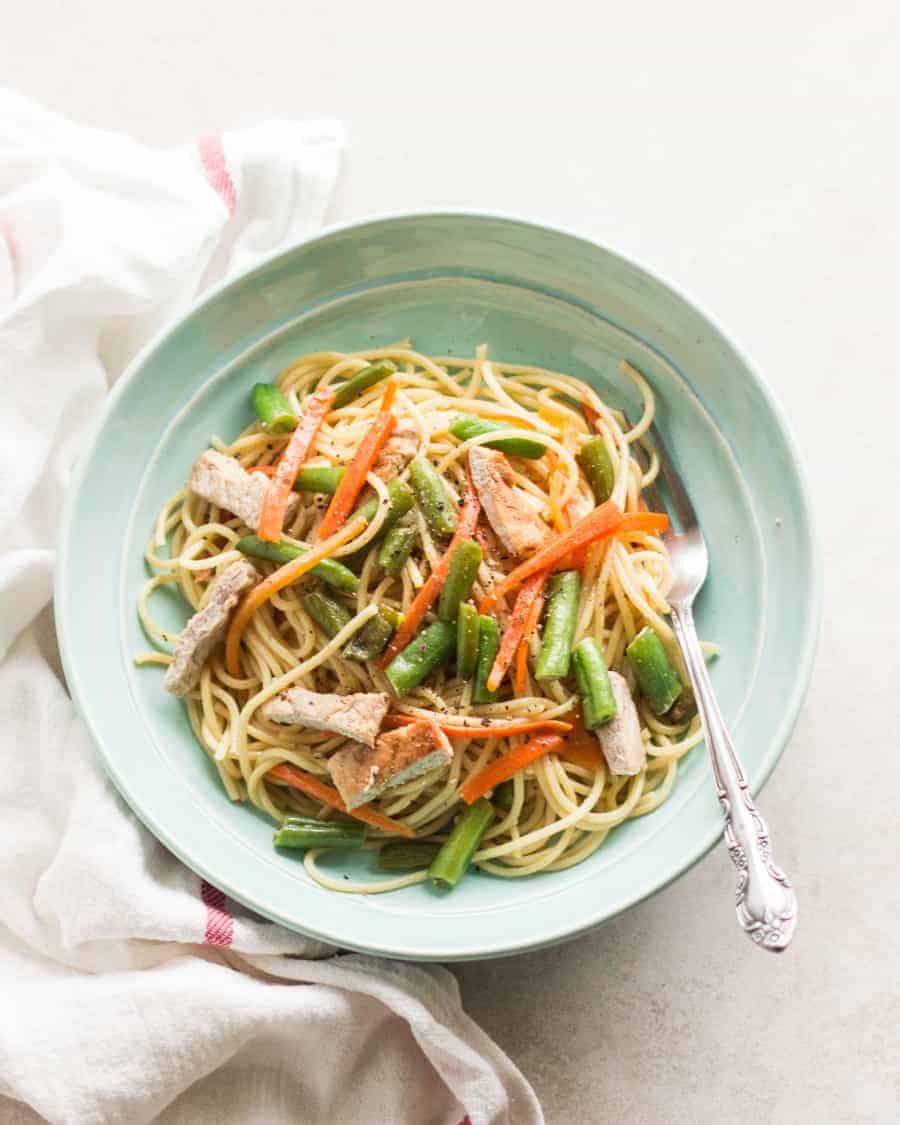 a big bowl of spaghetti noodles with green beans, carrots, pork and fork with white and red striped napkin on the side