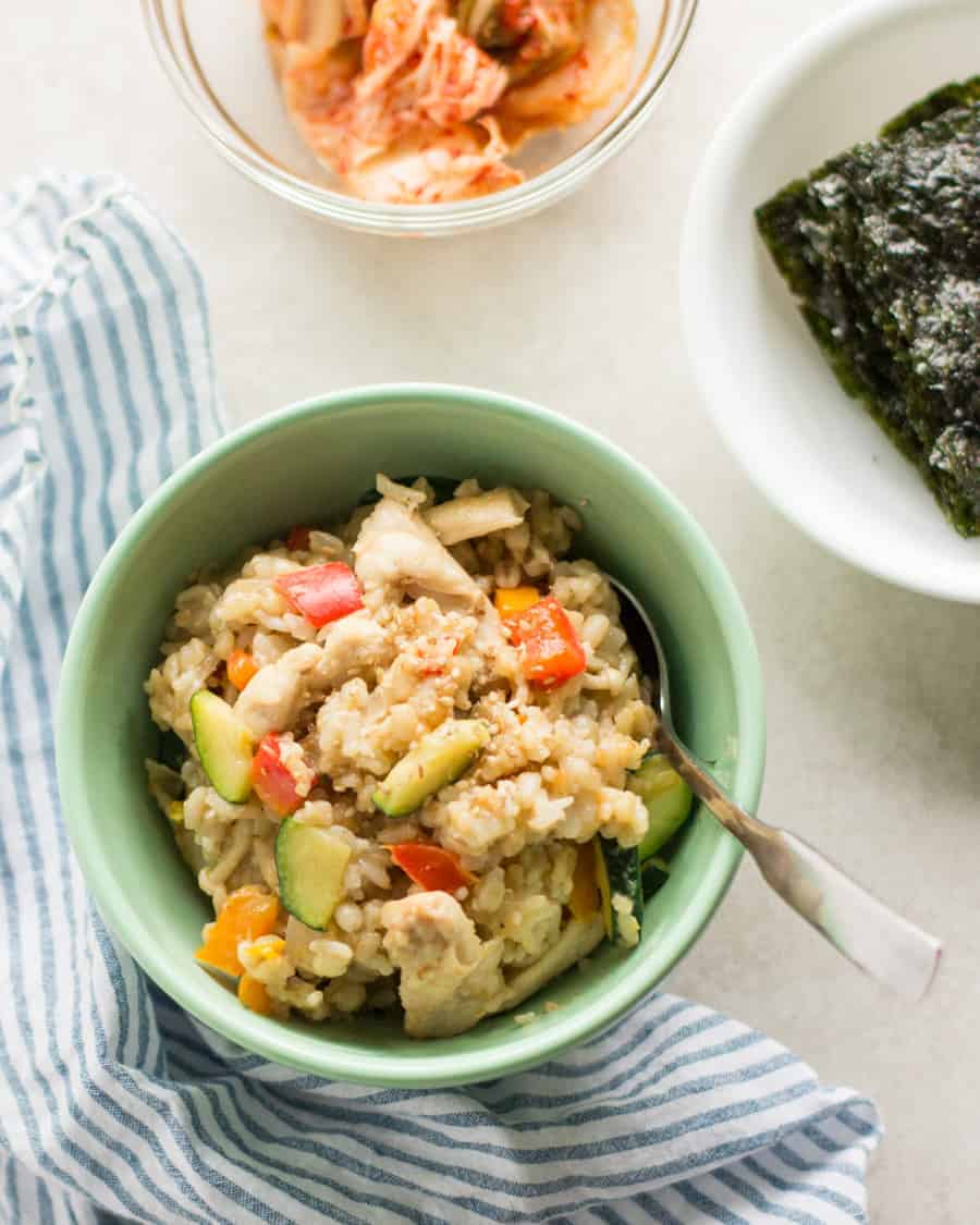fried rice in a green bowl with spoon laid on top of blue and white kitchen towel with kimchi and seaweed served in separate bowls