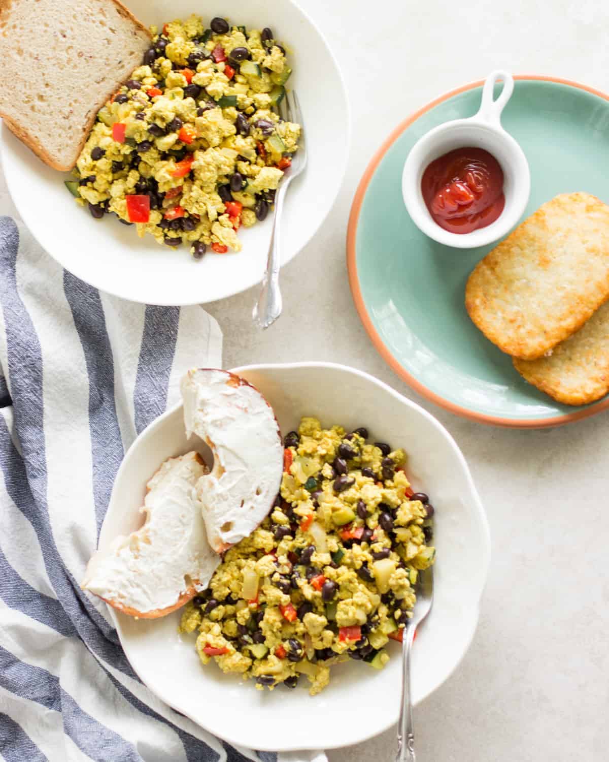tofu veggie scramble plated in two white bowls with toast, bagel, and hash browns