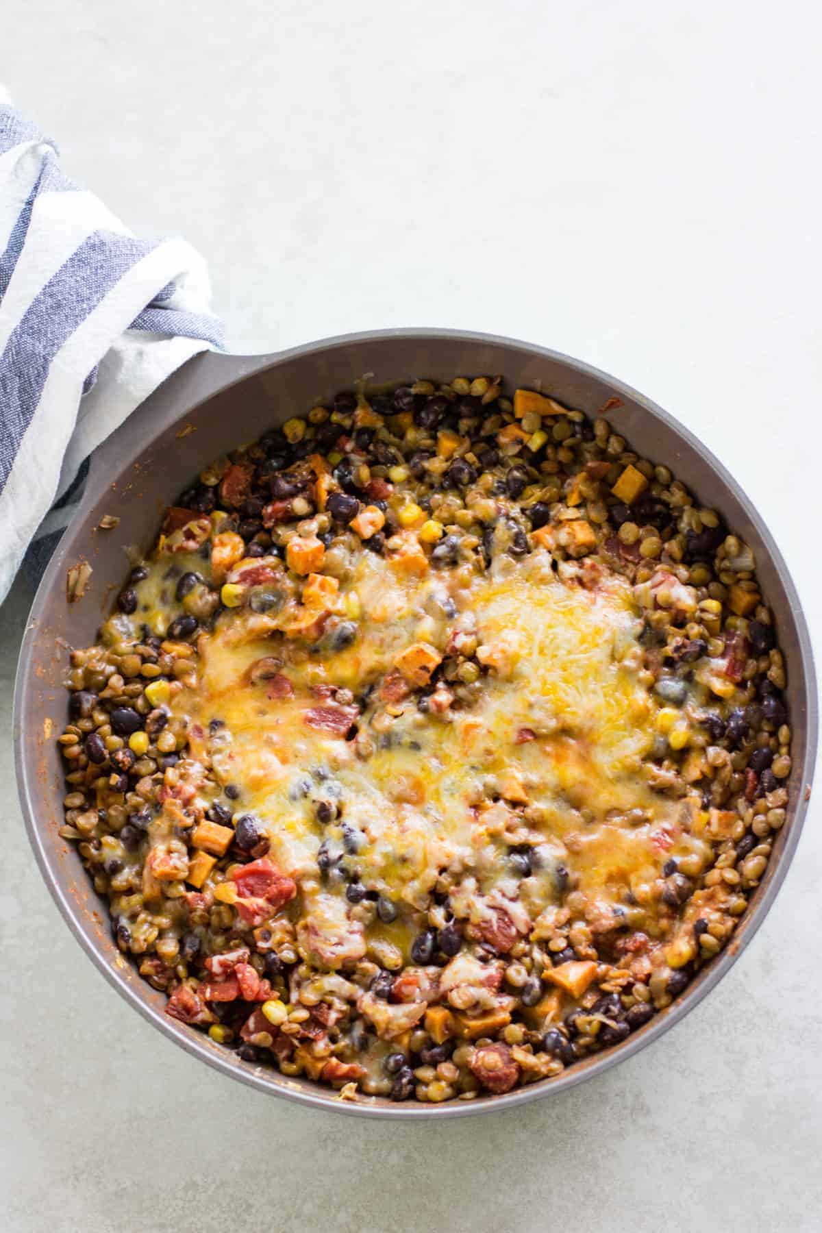 Mexican lentils in a large skillet topped with cheese and handle wrapped in blue and white striped kitchen towel