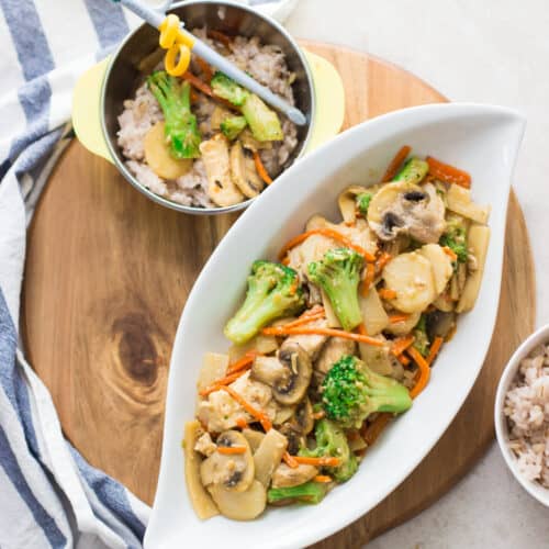 Chinese mushroom chicken plated on a large oval white dish and a bowl with rice and a small amount of the stir fry with chopsticks