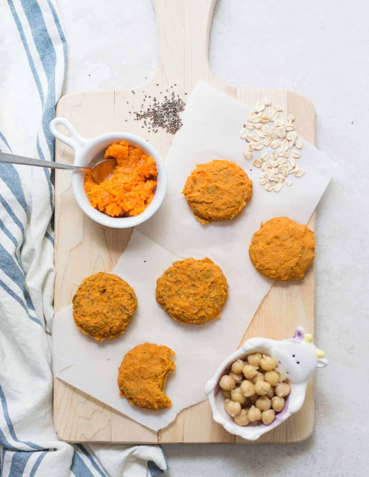 five baked cookies on a wooden board with mashed sweet potatoes, chickpeas, oats, and chia seeds