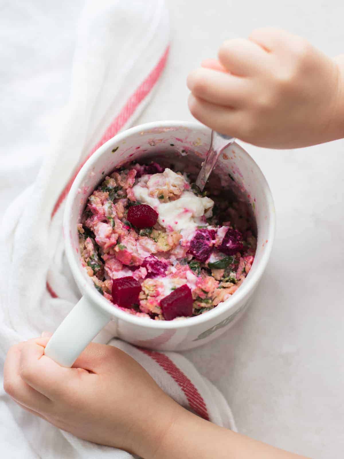 oatmeal topped with ricotta and beets in a mug with a toddler hand scooping with a spoon