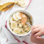 peanut butter banana oatmeal in a white mug with a toddler hand scooping with a spoon