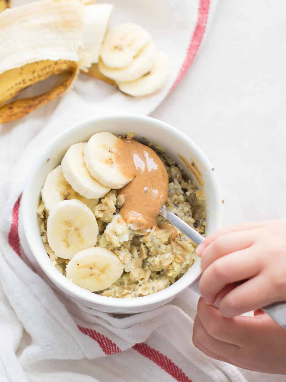 peanut butter banana oatmeal in a white mug with a toddler hand scooping with a spoon