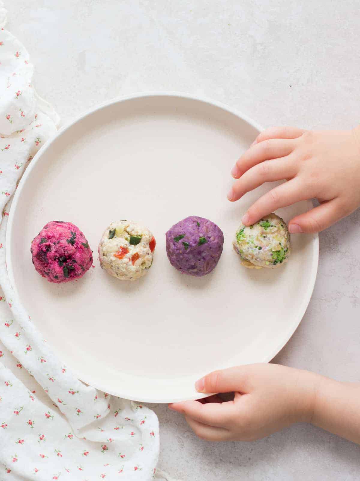 four oatmeal with vegetables rolled into balls with a baby's hand touching one.