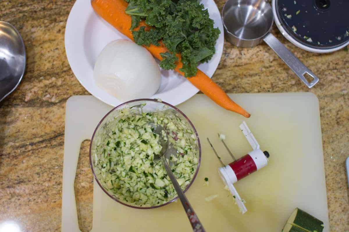 zucchini finely chopped in a mini manual food chopper with carrots and kale in the background