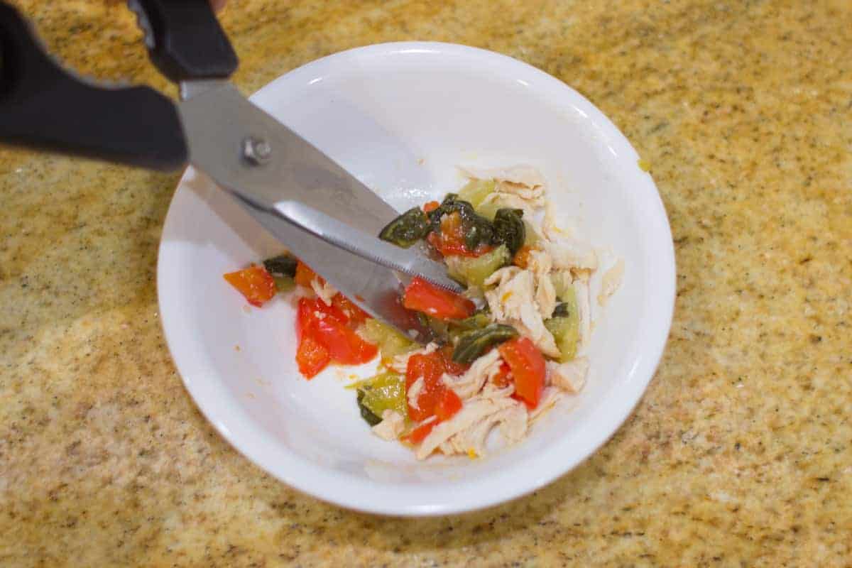 cooked chicken and vegetables being chopped with kitchen scissors in a white bowl