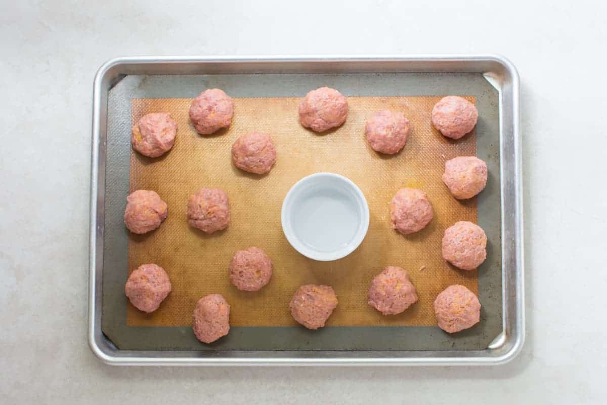 uncooked baby meatballs on a baking sheet pan with an oven safe dish filled with water in the center