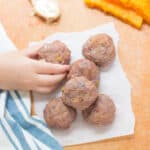 a close up shot of 6 baby meatballs on a wooden board with toddler's hand grabbing one