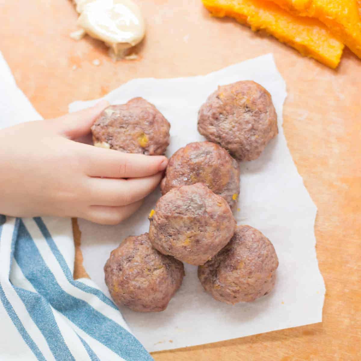 a close up shot of 6 baby meatballs on a wooden board with toddler's hand grabbing one