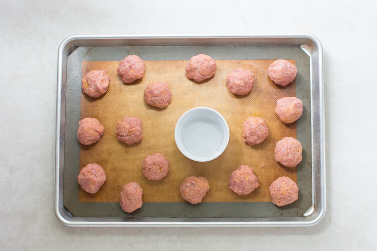 A small oven shaped dish filled with water placed in the middle of sheet pan with meatballs.