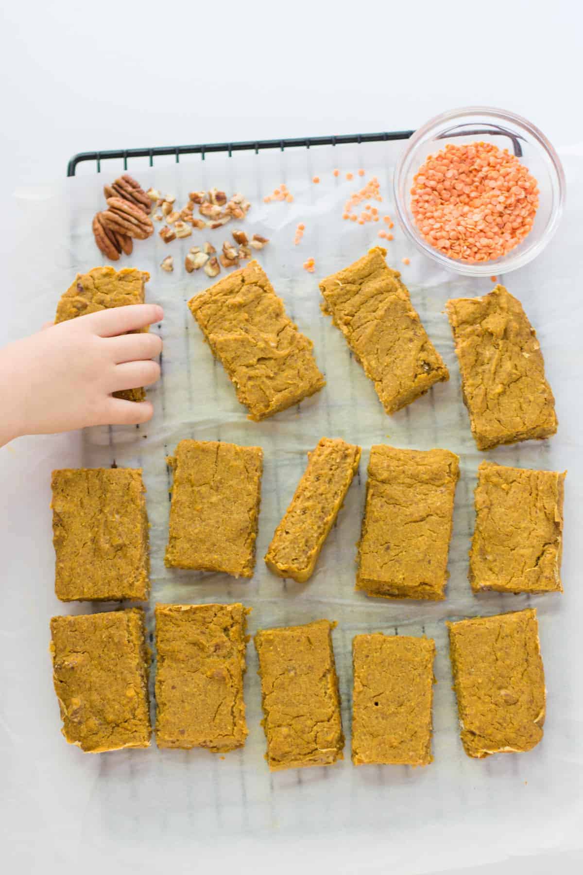 sliced pumpkin oatmeal bars on cooling rack with lentils and chopped pecans with toddler's hand reaching for one piece
