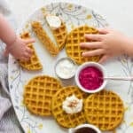 A large plate with cooked waffles, toppings, and little hands grabbing them.
