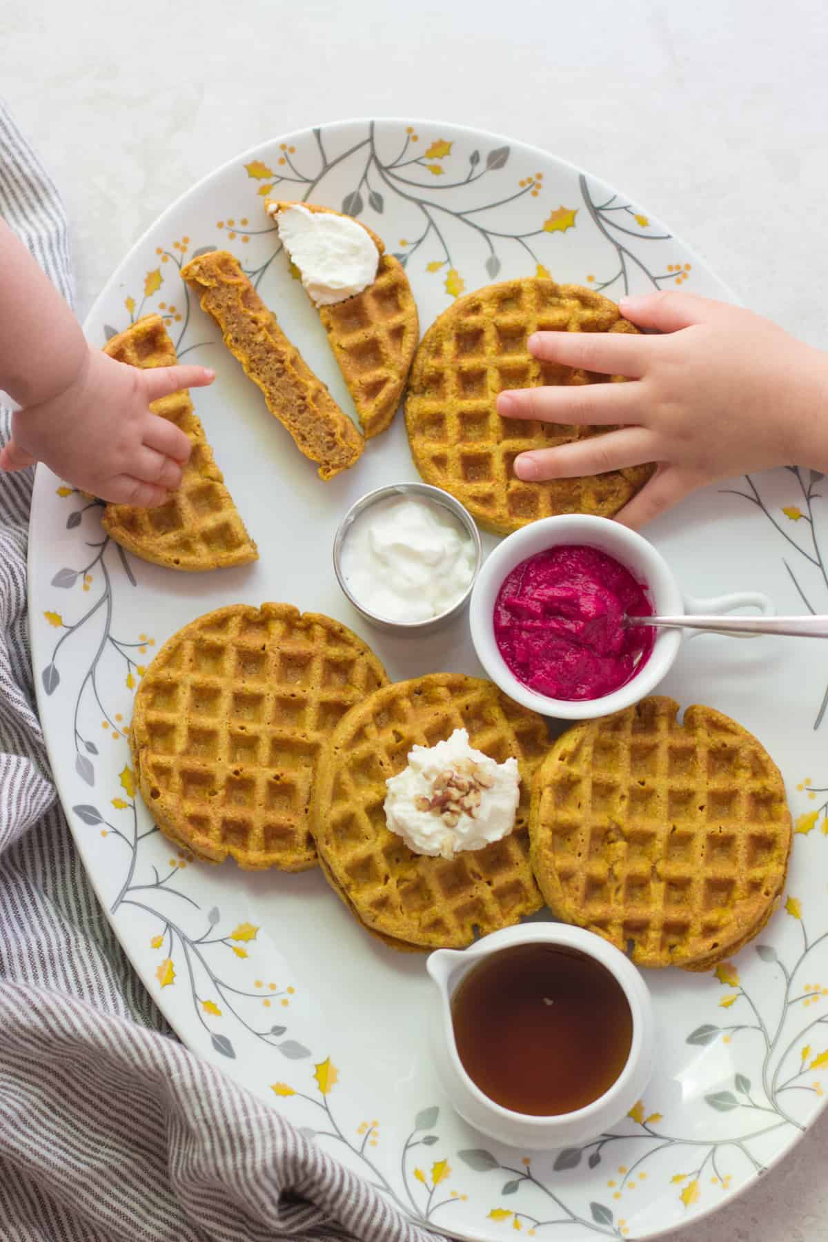 A large plate with cooked waffles, toppings, and little hands grabbing them.