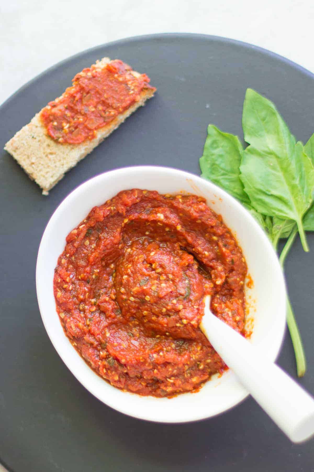 baby-friendly pizza sauce in a white bowl with a spoon and above it a toast spread with sauce and fresh basil on the side