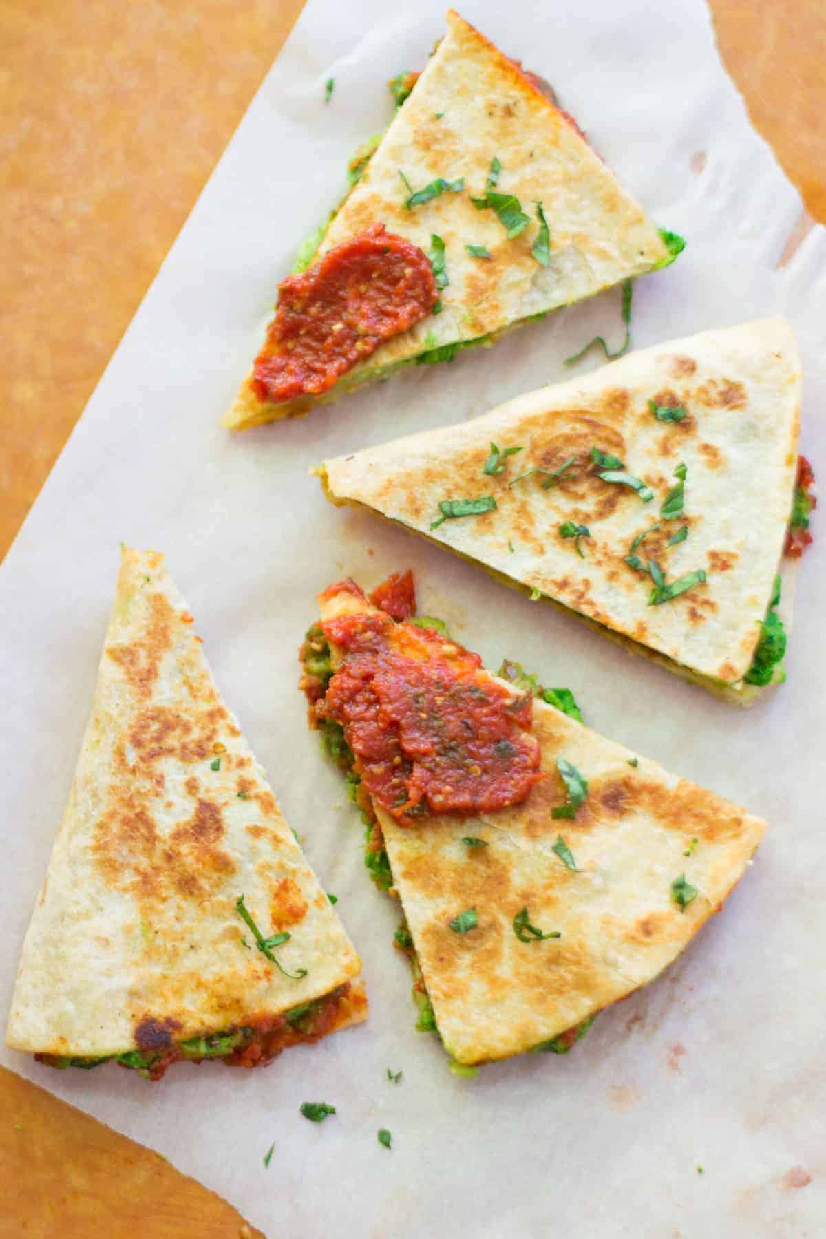 four pizza quesadilla triangles spread out on a parchment paper with pizza sauce spread on two pieces and fresh basil sprinkled throughout