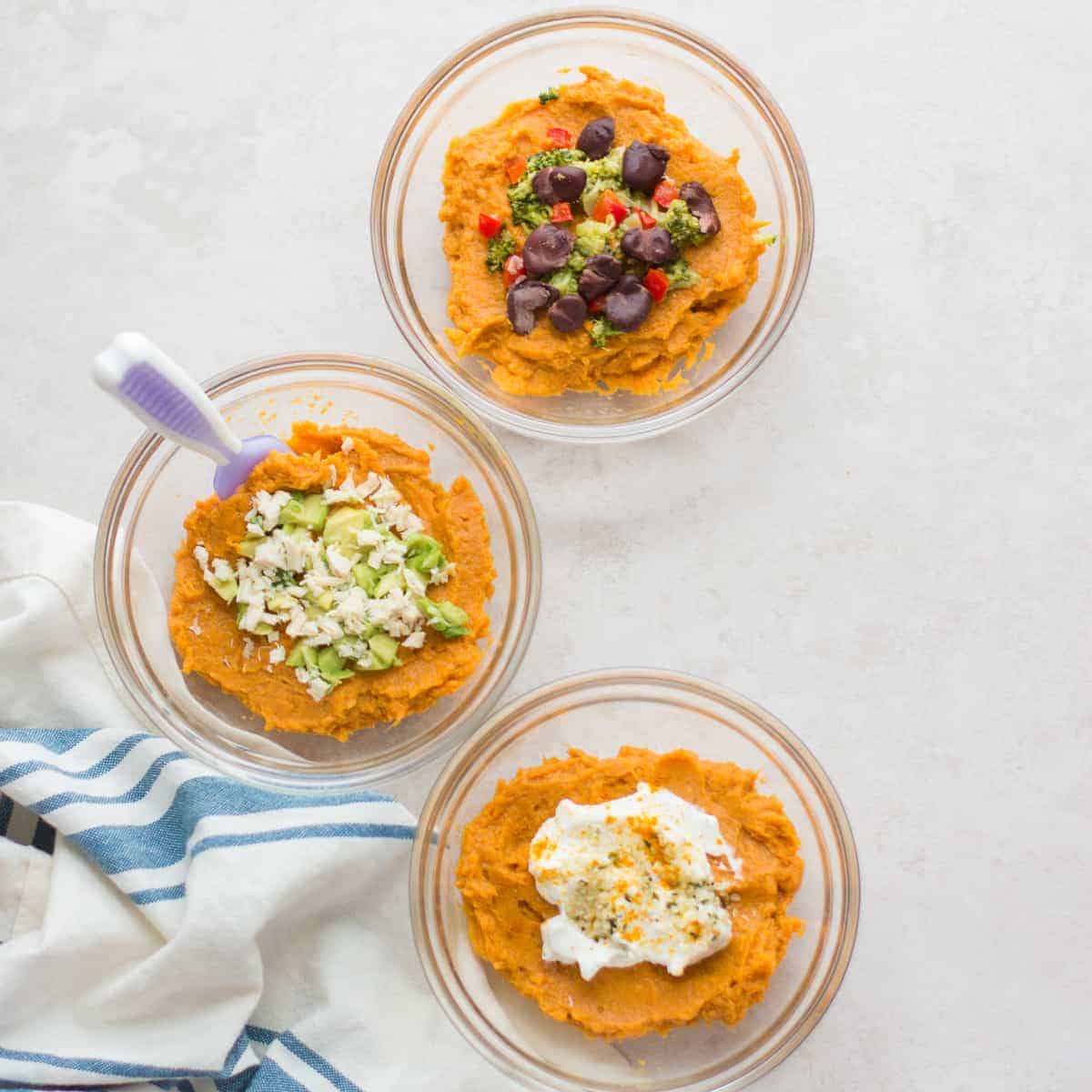 three different versions of mashed sweet potatoes in separate glass bowls. One with black beans and broccoli, second one with chicken and avocado, and third one with yogurt, hemp seeds, and turmeric