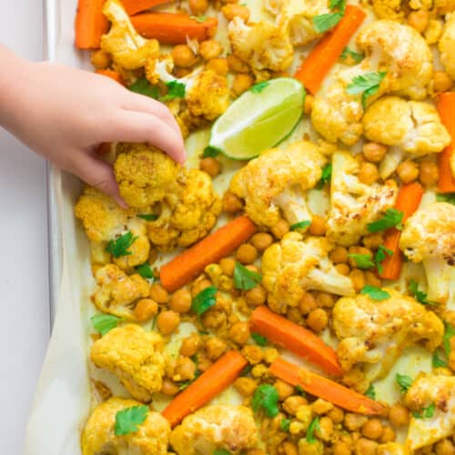 indian spiced roasted cauliflower and chickpeas on a baking sheet lined with parchment paper and toddler grabbing one cauliflower