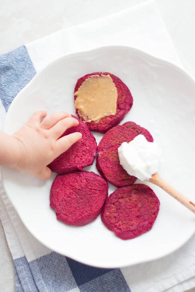 five cooked beet pancakes with yogurt and peanut butter spread on top of two and a baby reaching for one pancake