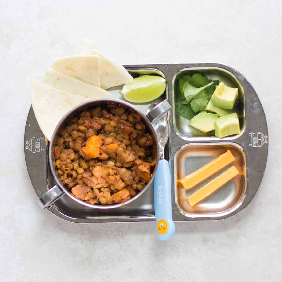 a toddler's bowl of lentil chili with tortilla, lime, avocado, cilantro, and cheese stick on the side