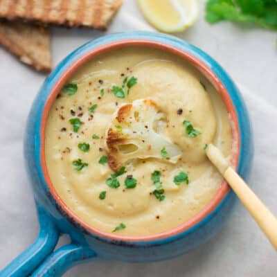 thick and creamy cauliflower chickpea soup in a blue soup bowl with one piece of roasted cauliflower and chopped cilantro with a gold spoon