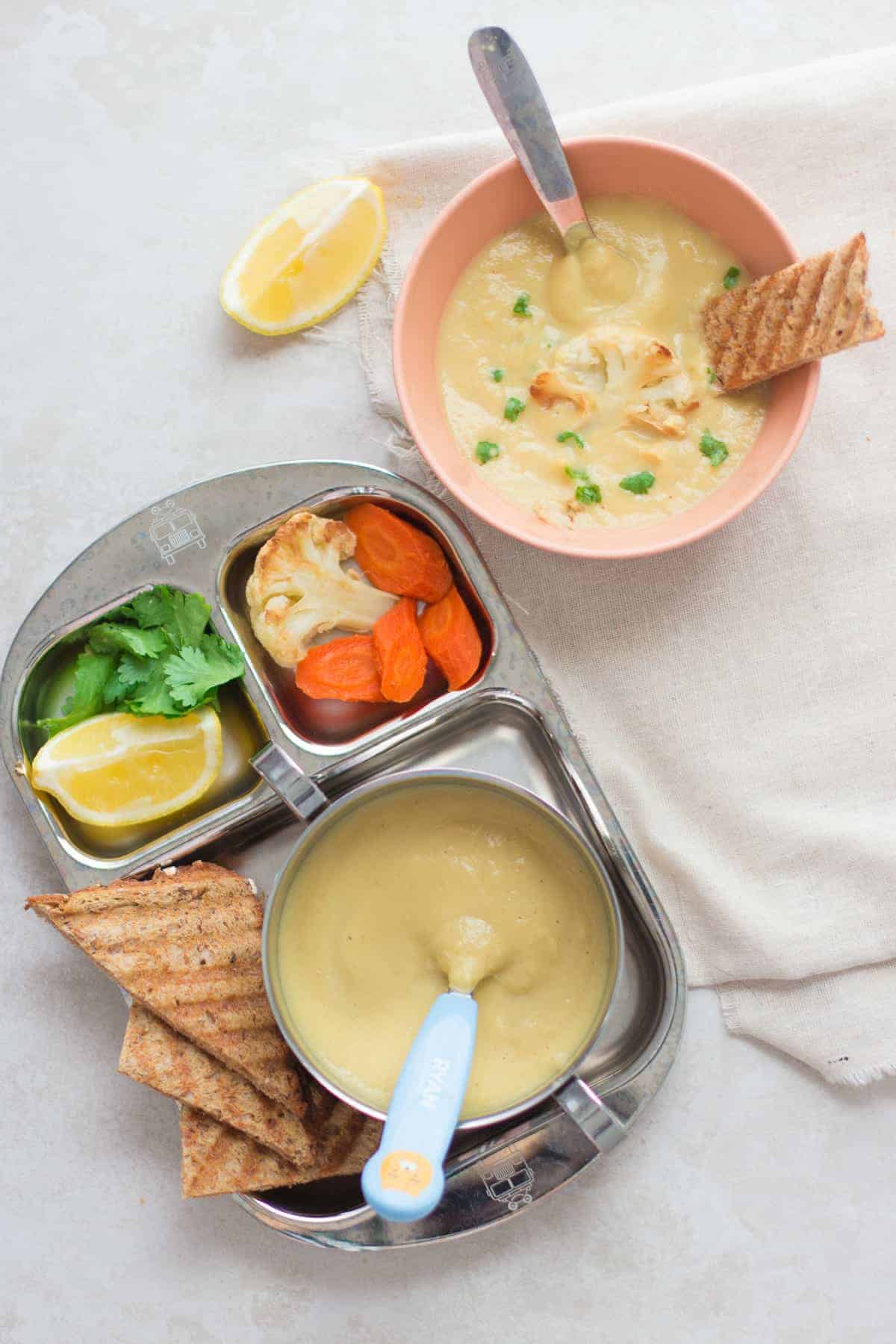 creamy chickpea soup served in a baby's bowl with a roasted cauliflower floret and chopped cilantro and toast strip as well as in a toddler bowl with toasted bread, cilantro, lemon wedge, roasted cauliflower floret and carrots