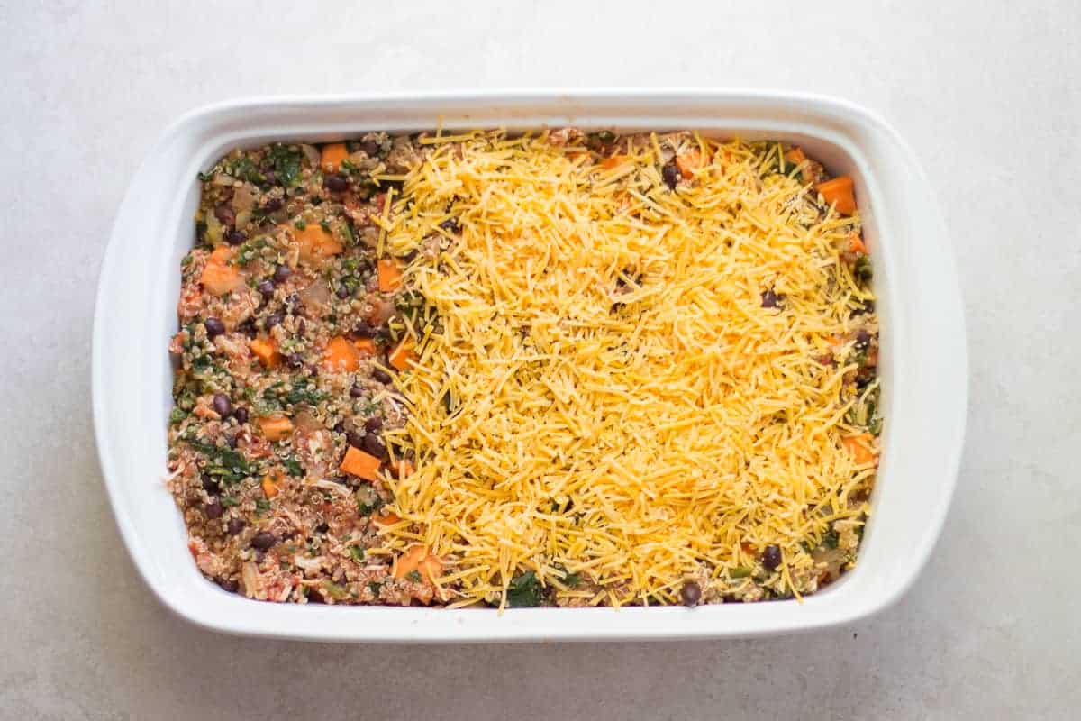 unbaked quinoa bake in a white baking dish with cheese spread on top of ¾ of the dish