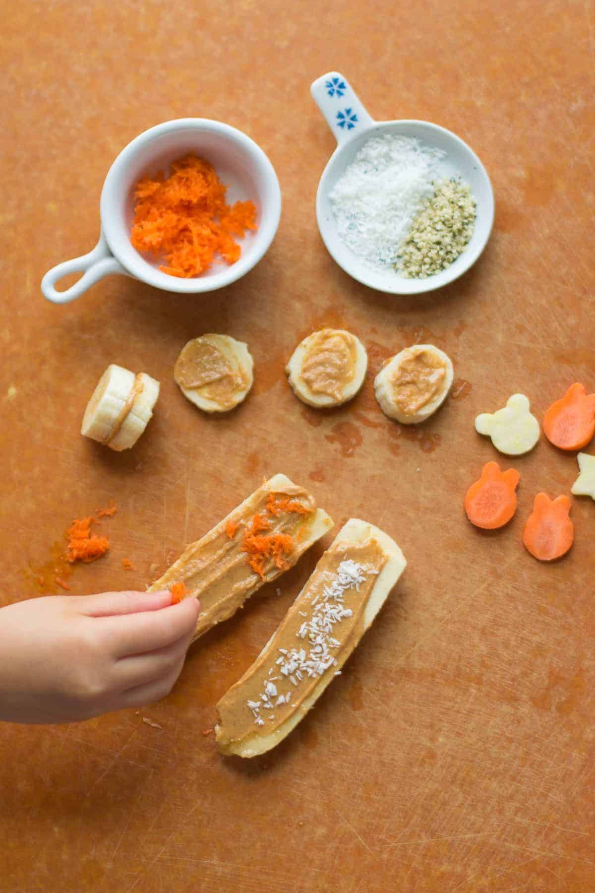 banana logs with peanut butter spread on top along with sliced bananas, grated carrots, coconut, hemp seeds, animal shaped raw zucchini and carrots on a wooden board.
