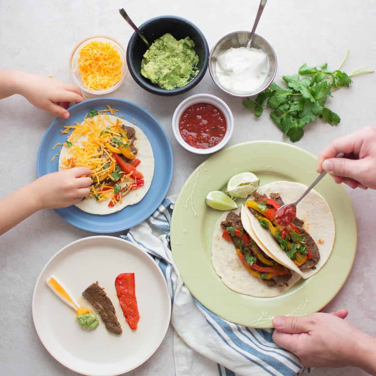 Steak fajitas served family-style with an assortment of toppings and toddler, baby, and adult's plate with tortilla.
