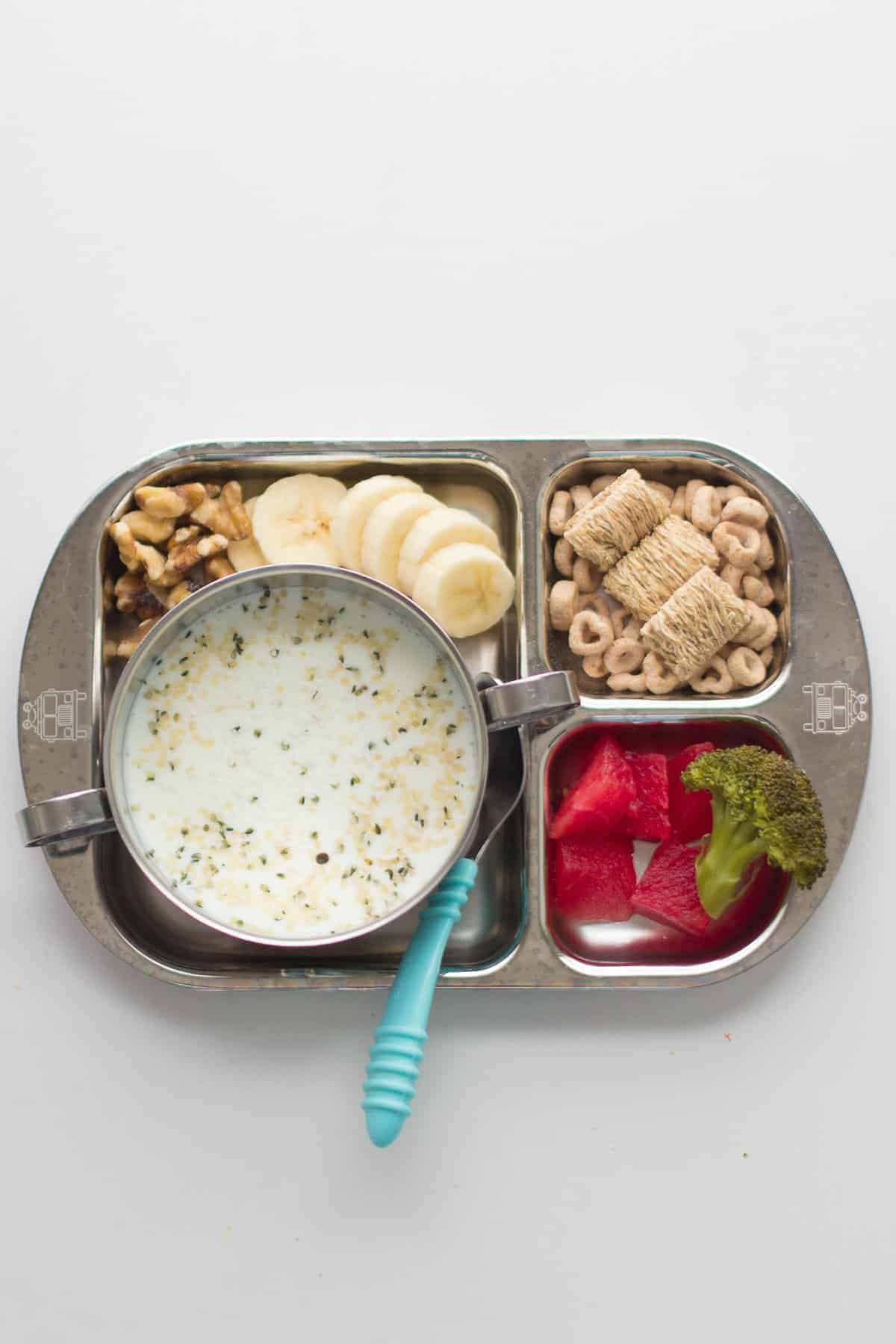 milk in a stainless steel bowl along with sliced bananas, walnuts, cereal, watermelon, and broccoli on a stainless steel plate.