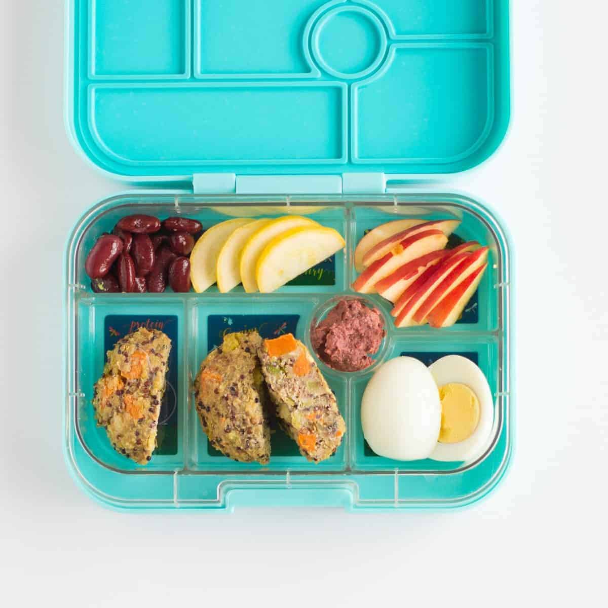 toddler lunchbox with quinoa cakes, hard boiled egg, sliced apples, zucchini, and red kidney beans.