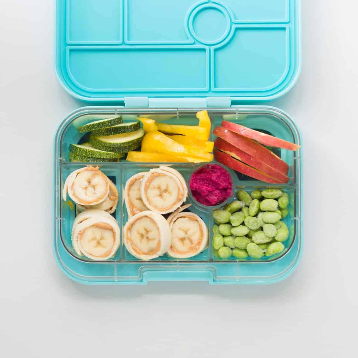 pb banana roll ups in a blue lunchbox with sliced zucchini, bell peppers, apple slices, edamame, and beet hummus.