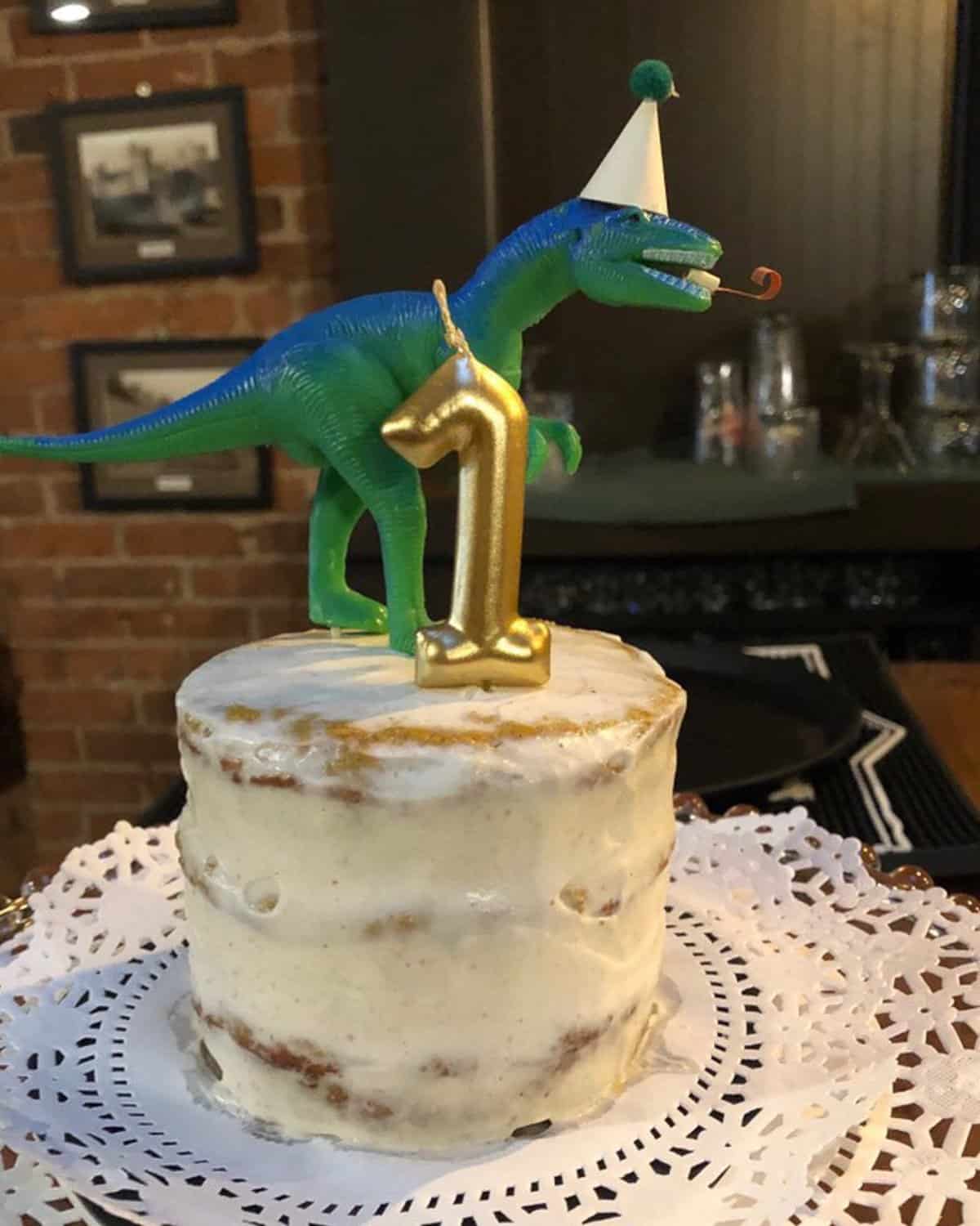 a three layered frosted cake with number one candle and a dinosaur.