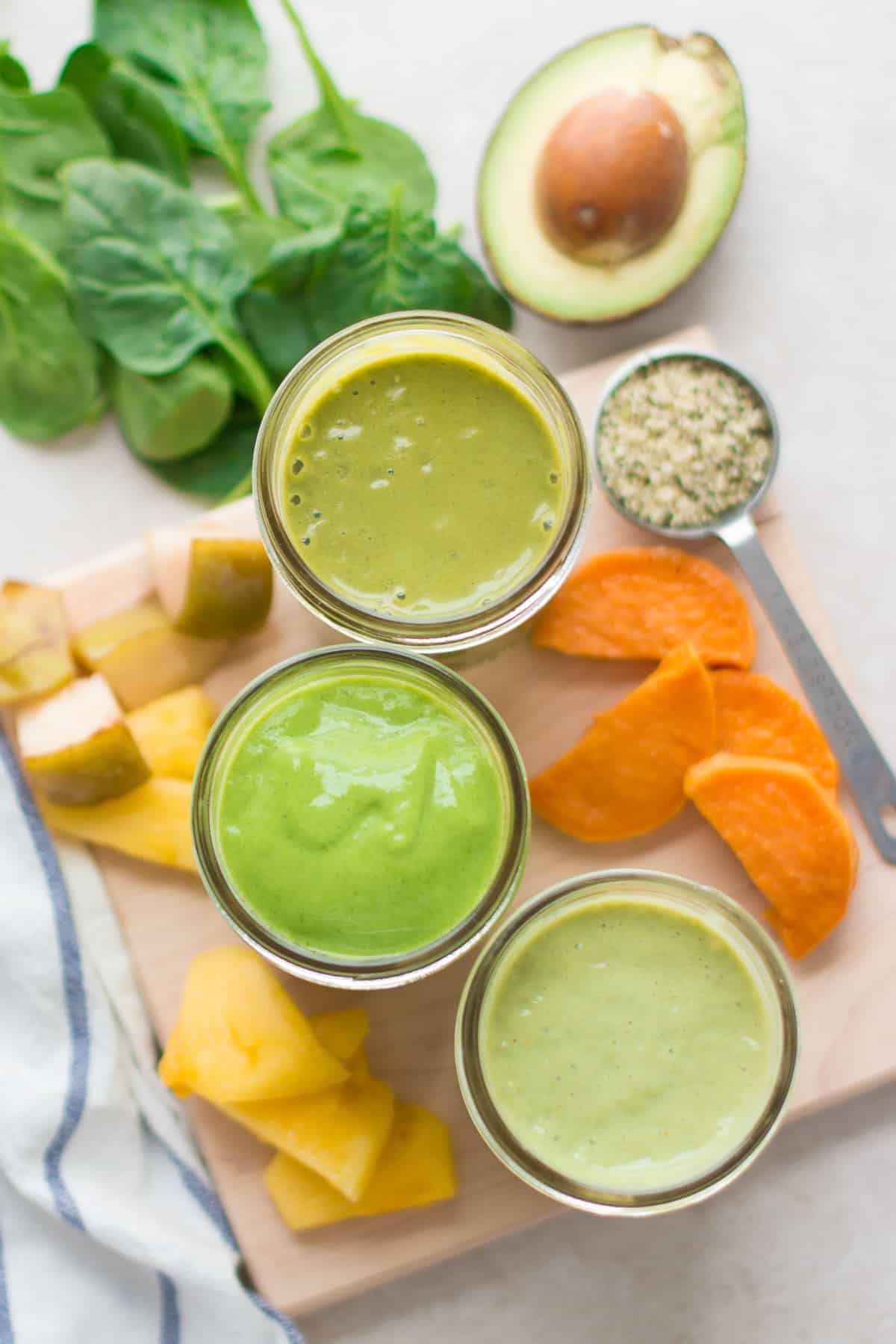 three green smoothies in glass jars for kids with various toppings surrounding them.