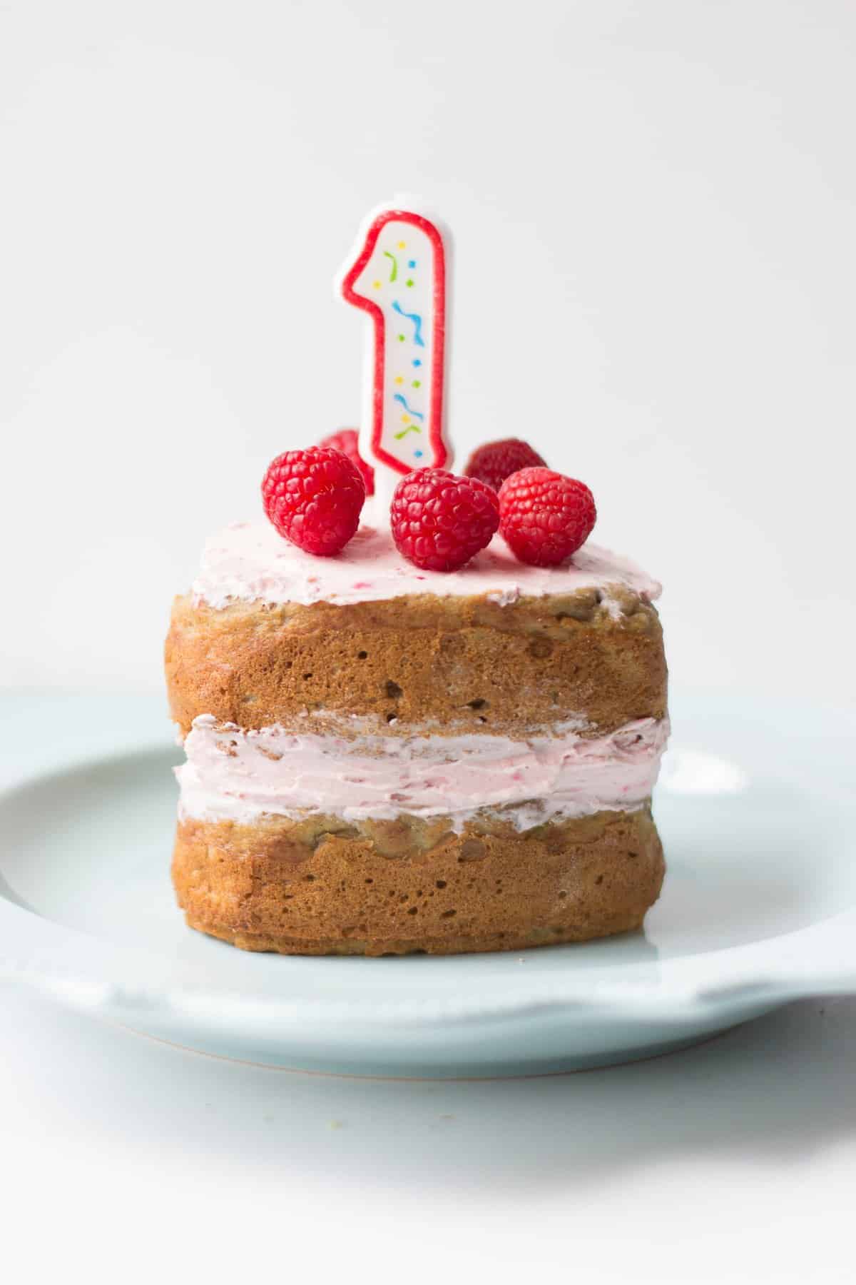 two layered birthday cake with cream cheese frosting in the middle and at the top with fresh raspberries and number 1 candle.