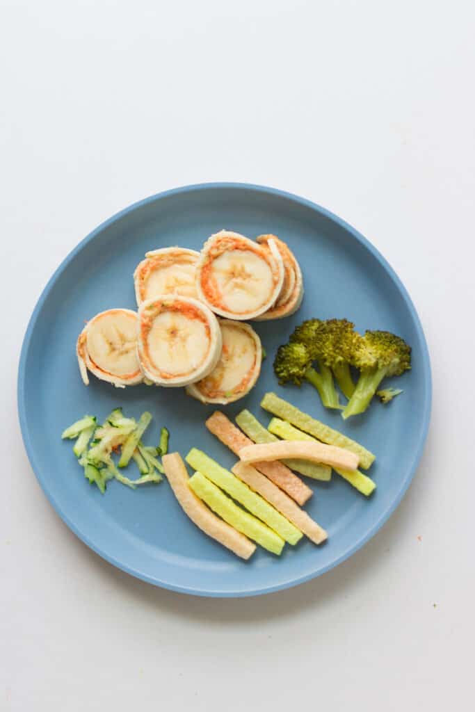 a toddler's lunch plate with peanut butter banana carrot roll ups, broccoli, veggie straws, and shredded zucchini.