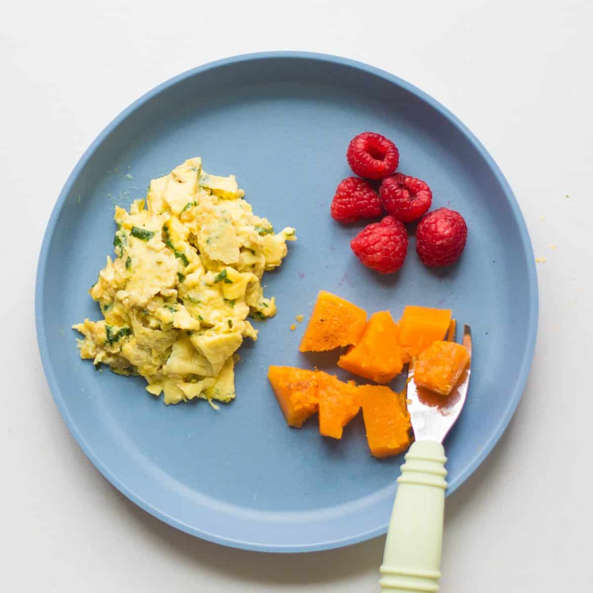 a toddler's breakfast plate with scrambled eggs, cubed butternut squash and raspberries on a blue plate.