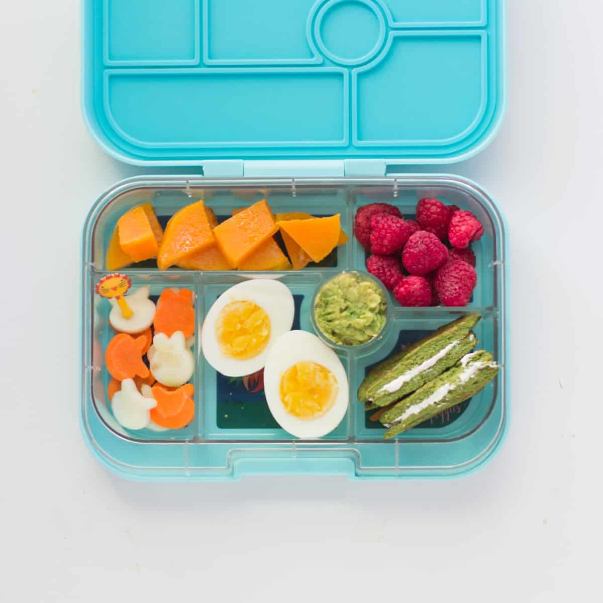 Spinach pancake sandwich with hard boiled egg, raspberries, butternut squash, hummus, and fun shaped zucchini and carrot pieces in a green lunchbox.