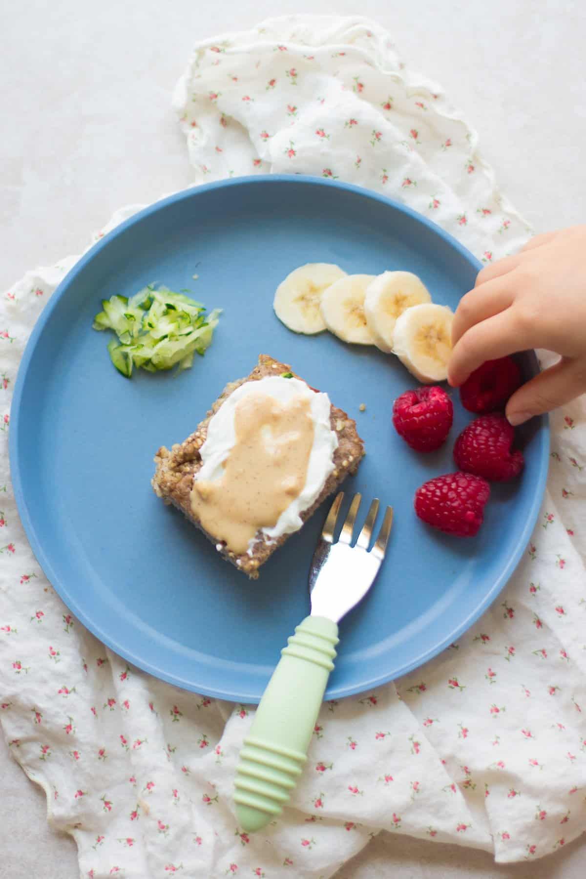 A slice of quinoa bake with yogurt and peanut butter with banana slices, raspberries, and grated zucchini on the side.