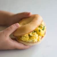 A toddler holding a mini bagel scrambled egg sandwich.