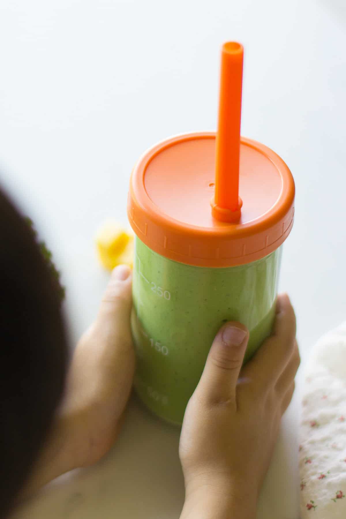 A toddler holding kale smoothie in a glass straw cup.