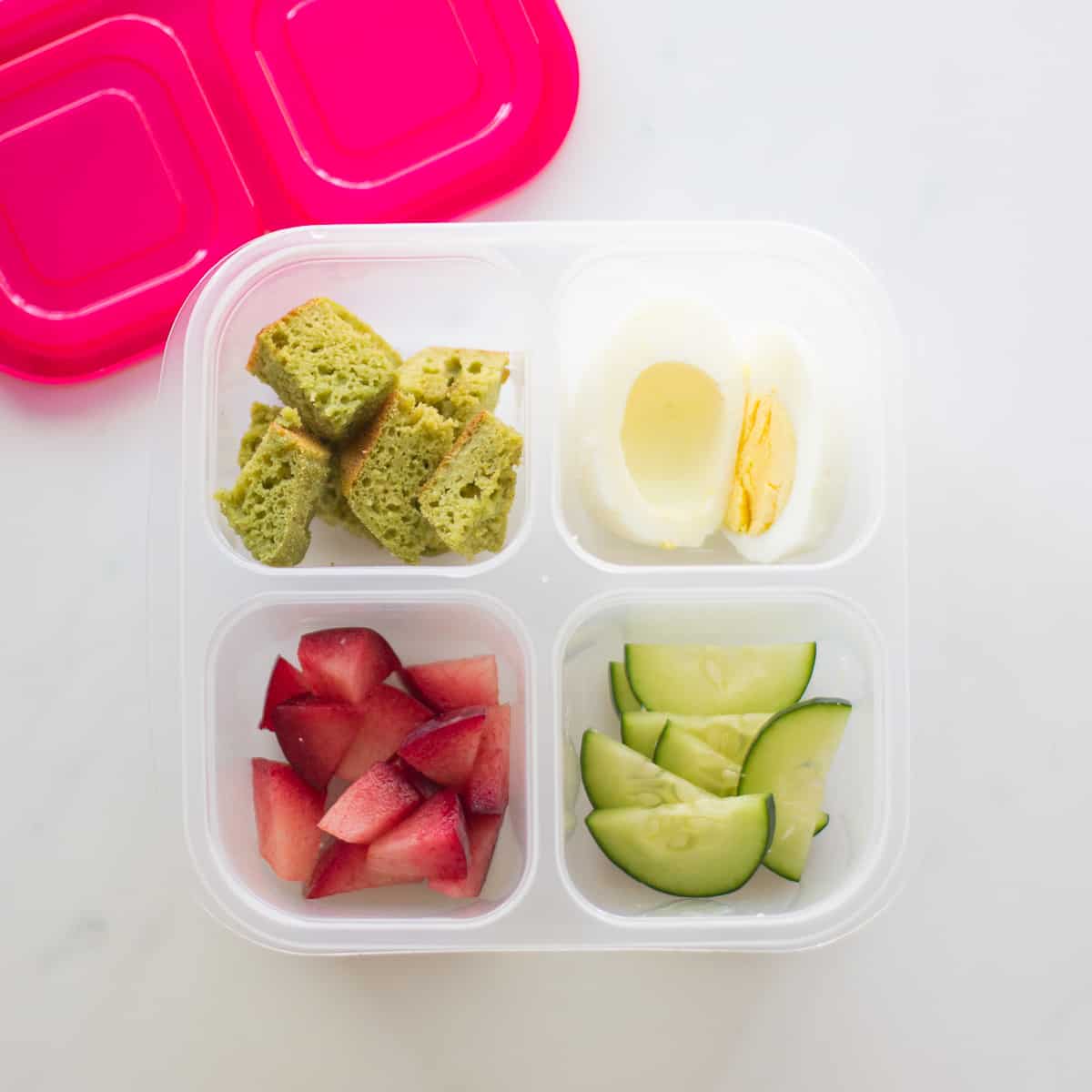 muffins, plums, cucumber, and hard boiled egg in plastic lunch box.